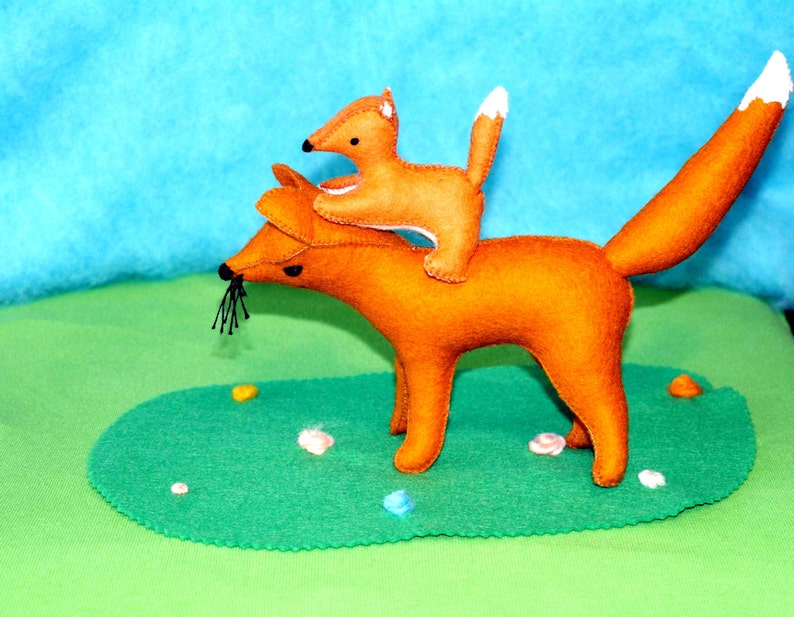 May include: A felt fox toy with a smaller fox riding on its back. The foxes are orange with white-tipped tails and black whiskers. They are set against a green and blue backdrop, creating a whimsical scene.