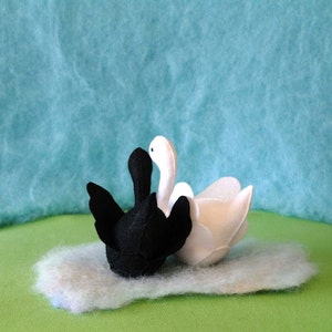 May include: Two felt swans, one black and one white, are nestled together on a fluffy white surface. The swans are facing each other, with their necks intertwined.