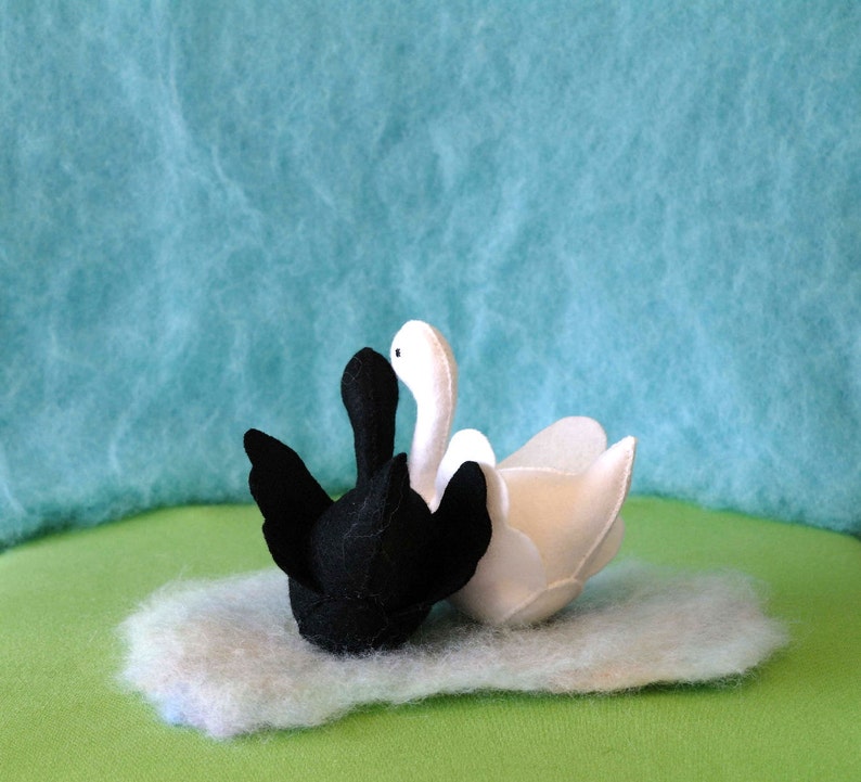 May include: Two felt swans, one black and one white, are nestled together on a fluffy white surface. The swans are facing each other, with their necks intertwined.