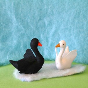 May include: A black swan and a white swan, both made of felt, are sitting on a green surface. The swans are facing each other.