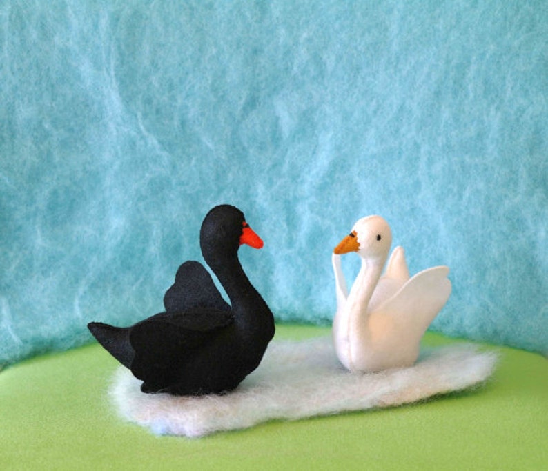 May include: A black swan and a white swan, both made of felt, are sitting on a green surface. The swans are facing each other.