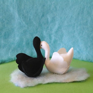 May include: A black swan and a white swan facing each other with their beaks touching. The swans are made of felt and are sitting on a fluffy white cloud.