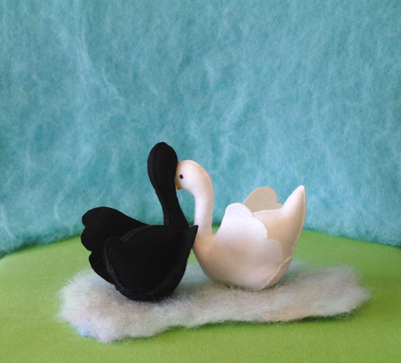 May include: A black swan and a white swan facing each other with their beaks touching. The swans are made of felt and are sitting on a fluffy white cloud.