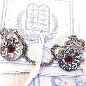 May include: Two silver-toned metal ornaments with a lion design and a red gemstone center. The ornaments are connected by a chain and rest on a white and blue patterned fabric. The lion is standing on a shield-shaped base.