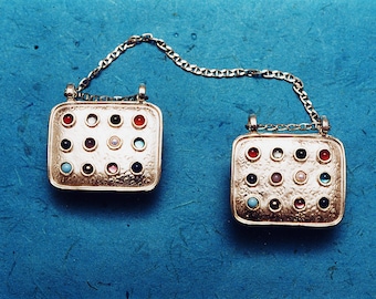 May include: Two gold pendants with a chain connecting them. Each pendant has a square shape with a textured surface and is adorned with 16 small, colorful gemstones.