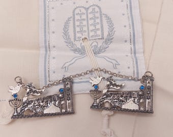 May include: A pair of silver-toned metal Torah breastplate ornaments featuring a menorah, a dove with an olive branch, and the word Peace. Each piece is accented with a small blue stone and connected by a silver chain, resting on a white fabric.