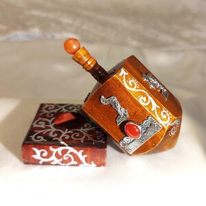 May include: A wooden dreidel featuring a polished brown finish, silver-toned filigree accents, and a small orange bead at the top. Beside it sits a square wooden base with matching silver-toned scrollwork and a circular cutout in the center.