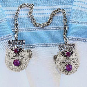 May include: Silver-toned tallit clips with a menorah design and a central star, each featuring a round, purple gemstone. The clips are connected by a chain and rest on a blue and white striped fabric.