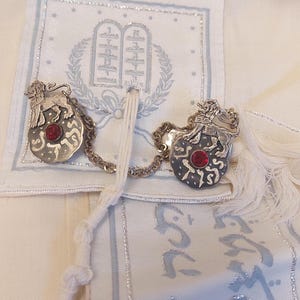 May include: Close-up of a white and light blue religious garment with silver embellishments. Two silver lion-shaped clasps with red gems are connected by a chain. The garment features a design of the Ten Commandments and Hebrew text.