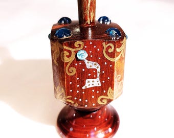 May include: A wooden dreidel featuring a rich brown finish with gold painted scrollwork and white polka dots. The top and sides are adorned with small blue faceted gems. A white Hebrew letter is painted on the front face of the spinning top.