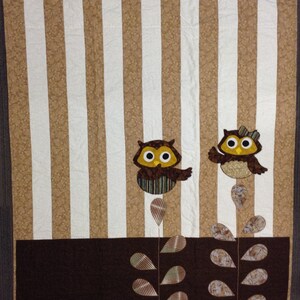 May include: A brown and white striped quilt with two brown owls perched on branches with green leaves. The owls have yellow eyes and brown beaks. The quilt has a brown border.