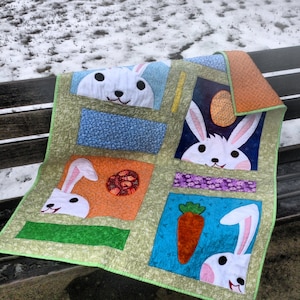May include: A green, orange, blue, and purple patchwork quilt with a white bunny rabbit design. The quilt features four bunny rabbits peeking out of different colored squares. The quilt is sitting on a wooden bench.
