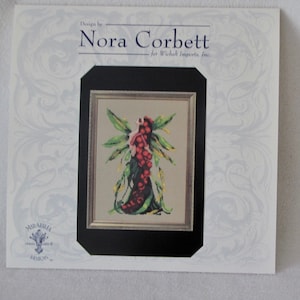 May include: Cross stitch pattern featuring a fairy with green wings and red berries. The pattern is framed in black and gold and has the text "Design by Nora Corbett for Wichelt Imports, Inc. Mirabilia Cross Stitch Designs."