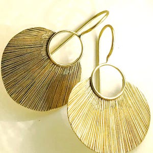 May include: A pair of gold-colored earrings. Each earring features a large, textured, fan-shaped design with radial lines, connected to a circular accent and a curved ear wire. The earrings have a bright, metallic finish.