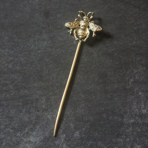 May include: A gold bee hair stick with a detailed bee design. The stick is thin and long, perfect for holding hair in place.