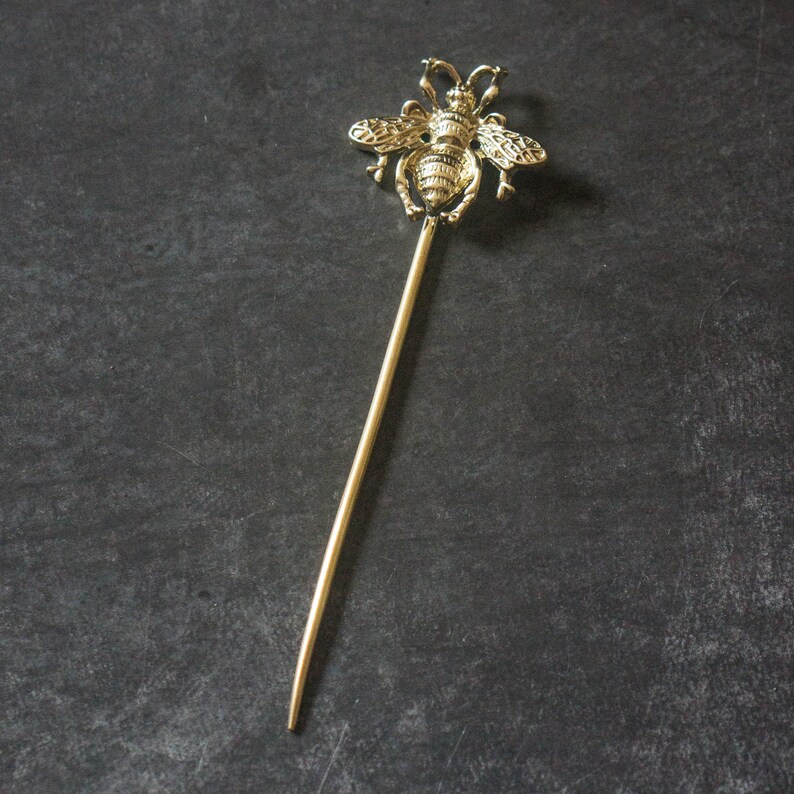 May include: A gold bee hair stick with a detailed bee design. The stick is thin and long, perfect for holding hair in place.