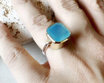 May include: A gold ring featuring a large, light blue gemstone. The ring is set in a gold band and has a square shape.