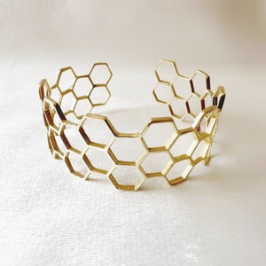 May include: A gold-coloured cuff bracelet with a honeycomb design. The openwork bracelet features a series of interconnected hexagonal shapes, creating a geometric pattern. The bracelet is made of a shiny, metallic material and is displayed on a white surface.