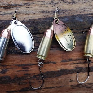 May include: Three fishing lures with bullet-shaped bodies and silver or gold-colored spinner blades. The lures have a hook at the bottom and are set against a wooden background. One lure has the text "French Spinner 3" on the blade.