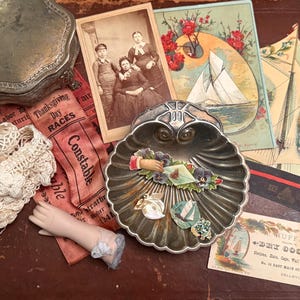 May include: An assortment of vintage items, including a silver trinket box, antique postcards with sailboat illustrations, a porcelain doll arm, and a decorative shell-shaped dish. Also includes a lace trim and a ribbon that says "Thanksgiving Day Races".