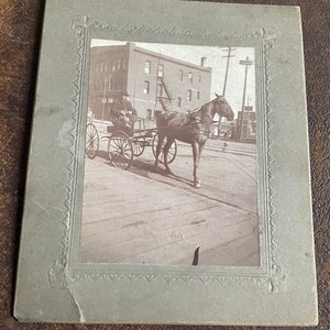 May include: A black and white photo of a horse-drawn carriage on a city street. The carriage is being driven by a person in a hat and coat. The horse is pulling the carriage on a cobblestone street. The photo is framed in a gray frame with a decorative border.