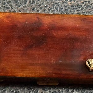 May include: A rectangular, wooden box with a rich, reddish-brown finish. A small, gold-colored crown emblem is affixed to the front. The box has a hinged lid and a clasp closure, suggesting it is designed for storage.