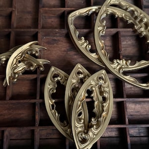 May include: A collection of gold-colored metal hardware pieces featuring intricate scrollwork and openwork designs. The set includes several pointed, leaf-shaped components and two larger, rounded oval frames, all displaying a metallic finish.