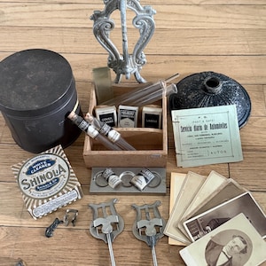 May include: A collection of vintage items including a black cylindrical tin, a wooden box with glass test tubes and Gilbert Chemistry labels, a silver-toned ornate stand, a Shinola White Cleaner box, metal music stand clips, and old photographs.