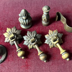 May include: An assortment of antique brass hardware components, including decorative drawer pulls with leaf-shaped backplates, a small finial, a hook, and a circular base. The pieces are arranged on a deep red surface.