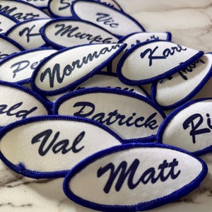 May include: A collection of oval-shaped white fabric patches with dark blue embroidered borders. Each patch features a unique name stitched in dark blue cursive script, including names such as Matt, Val, Rick, Patrick, Ronald, Norman, Karl, and Lloyd.