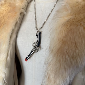 May include: A silver-toned necklace with a pendant shaped like a gun. The gun pendant is black with silver accents and a red tip. The necklace is displayed on a mannequin wearing a cream-colored jacket with a fur collar.