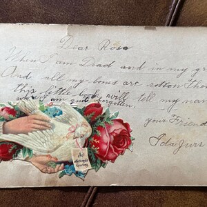 May include: A vintage postcard featuring handwritten cursive text that reads Dear Rose, When I am Dead and in my grave, And all my bones are rotten then, this little book will tell my name, your Friend, Ida Jurs. Includes a die-cut illustration of hands holding a white dove and red roses with a tag saying An affectionate Greeting.