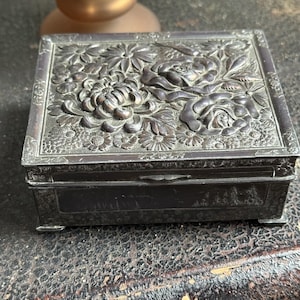 May include: A rectangular, antique silver-toned box with a hinged lid. The lid is intricately decorated with raised floral designs, including roses and chrysanthemums. The box has small feet and a dark, aged appearance, suggesting it is vintage.