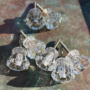 May include: A collection of six clear glass cabinet knobs featuring a faceted, multi-sided design. Each knob has a central metallic screw post extending from the back, with a circular metal base plate connecting the glass to the hardware.