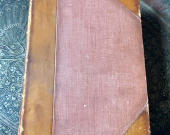 May include: A vintage book with a brown leather spine and a red cloth cover. The book has a worn, aged look and is likely a classic or antique.