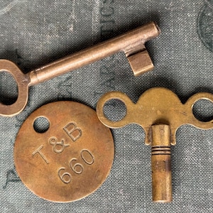 May include: An assortment of antique brass keys and a tag. The key on top has a rectangular head and a circular handle. The tag is round with the letters "T & B" and the number "660" engraved on it. The other key has a double-loop handle.