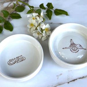 May include: Two white ceramic bowls with a brown design and text. One bowl has a sphinx design and the text "MADE IN GERMANY". The other bowl has the text "MADE IN GERMANY F.W. DEVOE & CO. NEW YORK".