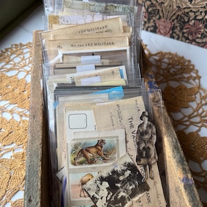 May include: A collection of vintage paper ephemera, including postcards, bank checks, and small illustrations of animals, housed in clear plastic sleeves within a weathered wooden box. The items feature printed text and black-and-white imagery.