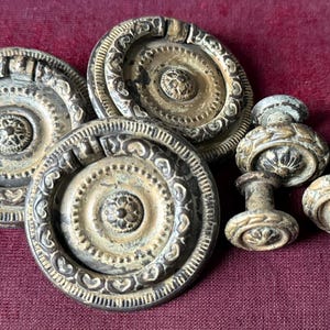 May include: Antique brass drawer pulls and knobs. The drawer pulls are circular with ornate detailing, while the knobs have a floral design. The hardware has a weathered appearance, suggesting age and use. The items are arranged on a burgundy surface.