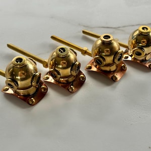 May include: Four brass cabinet knobs shaped like vintage deep-sea diving helmets. Each knob features a polished gold-tone spherical helmet with small circular portholes, attached to a copper-colored base plate and a long threaded metal bolt.