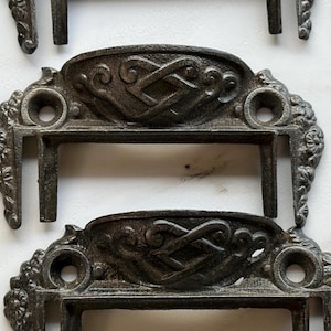 May include: A set of dark gray metal drawer pulls featuring an intricate raised knotwork pattern on the top arch and floral detailing on the side mounting plates. Each pull includes two circular screw holes for attachment to furniture surfaces.