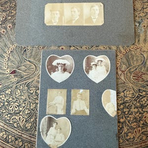 May include: Antique photo album with sepia-toned photographs. The album contains portraits of individuals and groups, some in heart-shaped frames. The album is gray and sits on a decorative surface with a gold and black pattern.