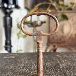 May include: Close-up of a vintage, rusty key. The key has an oval-shaped handle and a long, cylindrical stem. The metal has a weathered, aged appearance, with shades of brown and orange. The key is likely for a cabinet or antique furniture.