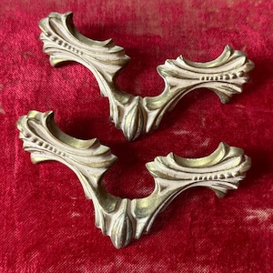 May include: Three antique, ornate drawer pulls with a cream and gold finish. The decorative hardware has a curved shape with detailed floral and beaded accents. The pulls are set against a red background.