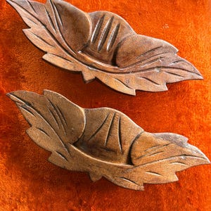 May include: Two brown wooden decorative objects shaped like stylized leaves. Each piece features carved vein details and a central raised section with vertical grooves, showcasing the natural wood grain and texture throughout the surface.