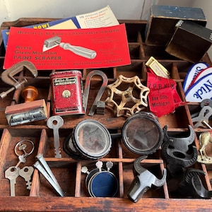 May include: A wooden printer's tray filled with various vintage items. Includes a red paint scraper advertisement, a red tin, keys, metal objects, a patch with "Nick" on it, and other small collectibles.