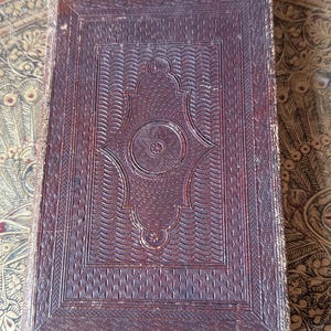 May include: A vintage, rectangular book with a dark brown, embossed leather cover. The cover features intricate patterns, including a central medallion and decorative borders. The edges show signs of wear, suggesting age and use.