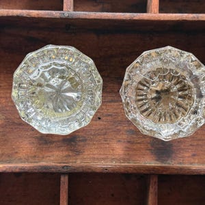 May include: Two clear glass door knobs with an octagonal shape and a faceted, starburst-like pattern radiating from the center. The knobs feature a reflective, transparent surface with intricate geometric detailing along the edges and a smooth top.