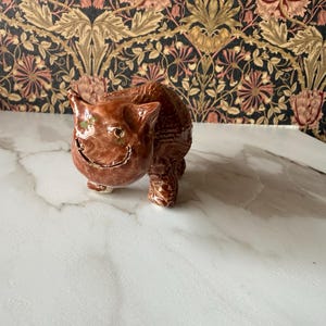 May include: A glossy brown ceramic figurine of a cat with pointed ears, a wide carved smile, and small green accents on the eyes. The cat is depicted in a standing position with a textured, rounded body and a smooth, reflective glaze finish.