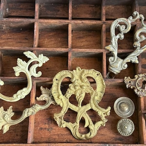 May include: A collection of antique brass hardware pieces, including ornate handles, keyhole covers, and decorative accents. The pieces are arranged in a wooden divided tray, showcasing their intricate designs and aged patina. The hardware is likely for furniture or doors.